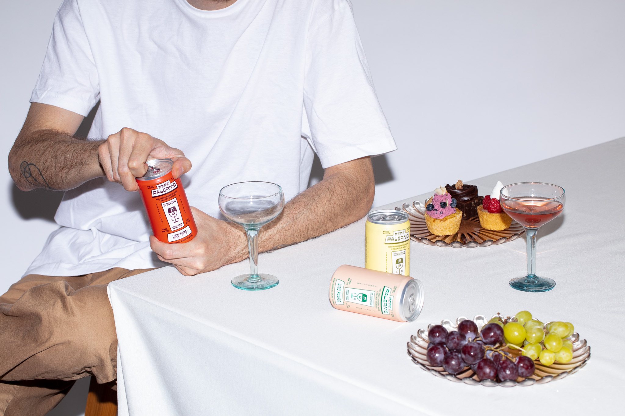 Young man opening a Pepe Palermo wine can, with wine glasses, cans, grapes, and pastries on a white table.